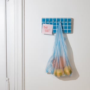 Puede incluir: Una bolsa de plástico azul llena de productos cuelga de un perchero de pared. El perchero está hecho de azulejos azules. Una nota adhesiva rosa con las palabras "Buy Eggs!" está adjunta.