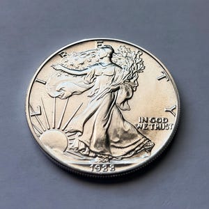 Puede incluir: Una moneda de plata con una mujer con un vestido fluido, sosteniendo ramas y caminando hacia el sol. La moneda tiene el texto "LIBERTY" e "IN GOD WE TRUST", con el año "1938" visible.
