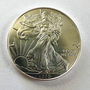 Może przedstawiać: Złota moneta przedstawiająca Lady Liberty idącą w kierunku słońca, odzianą w powłóczystą szatę, niosącą gałęzie. Moneta zawiera napis "IN GOD WE TRUST" oraz rok "2020".