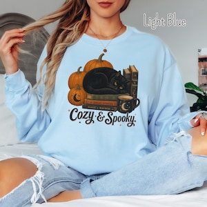 May include: A light blue crewneck sweatshirt featuring a graphic print of a black cat resting on a stack of books next to orange pumpkins and a dark mug. The text "Cozy & Spooky" is printed below the illustration in a dark brown script font.