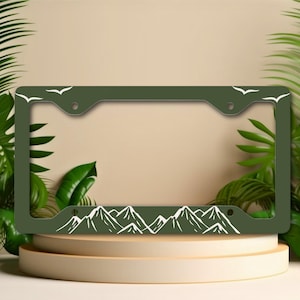 May include: A dark green license plate frame featuring a white mountain range design along the bottom and white bird silhouettes at the top corners. The frame is rectangular with pre-drilled holes for mounting. The design is simple and nature-themed.