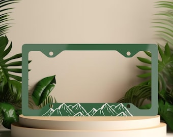 May include: A rectangular forest green license plate frame featuring a white line art graphic of a mountain range along the bottom edge. The frame includes four circular mounting holes, two at the top and two at the bottom, with a smooth finish.