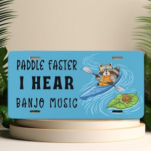 May include: A light blue license plate with the text "Paddle Faster I Hear Banjo Music." The plate features a cartoon raccoon in a kayak, wearing goggles and a life vest, paddling in blue water. A turtle rests on a lily pad.