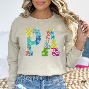 May include: Beige sweatshirt with the letters "PA" in a patchwork design. The letters are filled with colorful floral and nautical patterns, including sailboats, palm trees, and flowers. The sweatshirt has long sleeves and a crew neck.