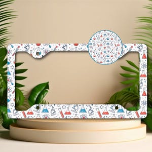 May include: White license plate frame with a repeating pattern of science-themed illustrations in red, blue, and black. The design includes beakers, microscopes, and atoms. A circular inset shows a close-up of the pattern.