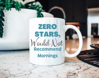 May include: White ceramic mug with the text "ZERO STARS, Would Not Recommend Mornings" in teal and black. The mug has a curved handle and is set on a marble surface. A white vase with green plants is in the background.