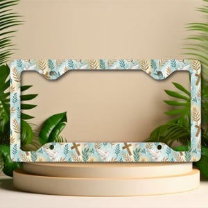 May include: License plate frame featuring a repeating pattern of crosses, doves, and leaves in blue, beige, and brown. The frame is rectangular with mounting cutouts. The design is set against a light blue background.