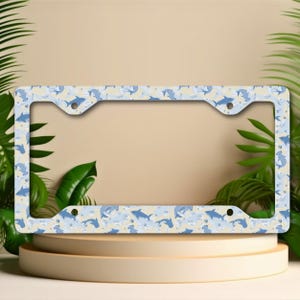 May include: A rectangular license plate frame featuring a repeating pattern of light blue and dark blue sharks on a pale cream background. The frame has four mounting holes, two at the top and two at the bottom, with a wide border design.