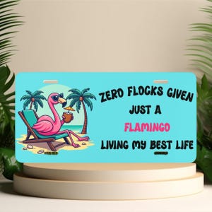 May include: A rectangular light blue license plate featuring a cartoon pink flamingo wearing sunglasses, lounging on a beach chair with a tropical drink. The text reads: ZERO FLOCKS GIVEN JUST A FLAMINGO LIVING MY BEST LIFE in black and pink lettering.