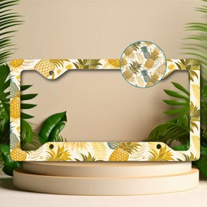 May include: License plate frame with a tropical pineapple pattern in yellow, gold, and green. The frame has a white background and a circular inset showing a close-up of the pineapple design. The frame is on a beige pedestal.