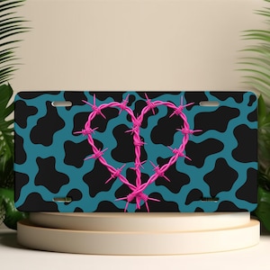 May include: A rectangular license plate featuring a teal and black cow print pattern across the background. Centered on the plate is a heart shape formed by bright pink barbed wire, complete with sharp spikes along the wire's length.
