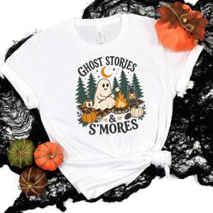 Op de afbeelding: Een wit t-shirt met een afbeelding van een geest die een marshmallow roostert boven een kampvuur, omringd door dennenbomen, pompoenen en een lantaarn. De tekst "GHOST STORIES & S'MORES" staat in een donker lettertype boven de afbeelding.