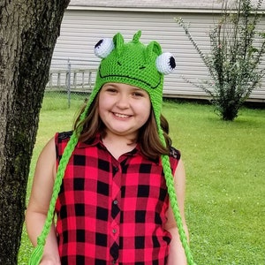 May include: A young person wearing a green crocheted frog hat with long green crocheted ties. The hat has white eyes and a black mouth. The person is wearing a red and black plaid shirt and black pants. They are standing in front of a tree.