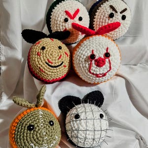 May include: Seven crocheted plush toys shaped like characters from horror movies. The toys are all different colors and have different facial expressions. They are arranged in a circle on a white crocheted surface.