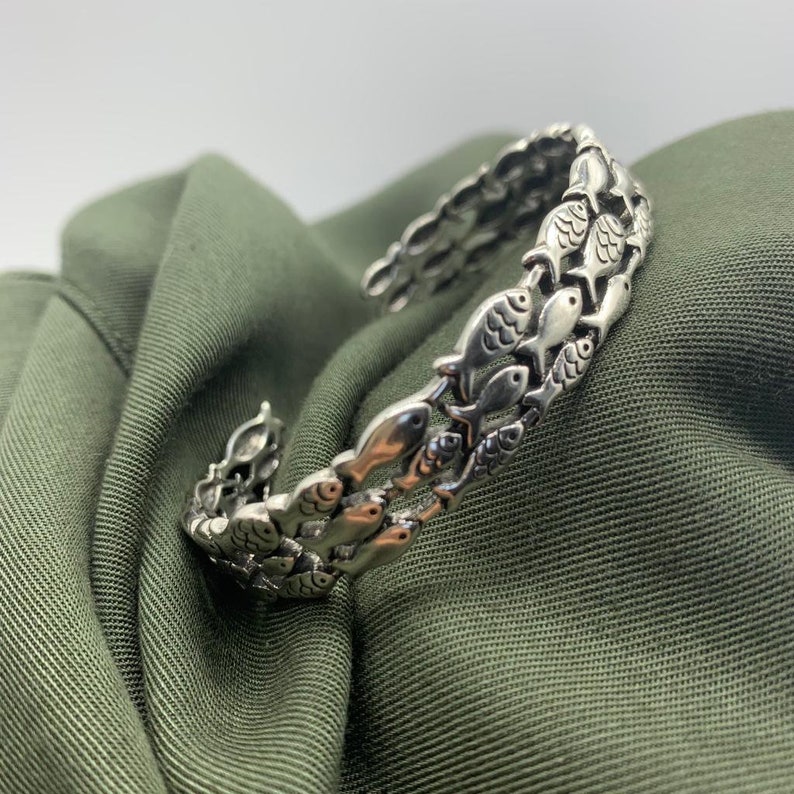 May include: A silver cuff bracelet featuring a repeating pattern of fish. The bracelet is open and flexible, with the fish arranged in rows. The bracelet is displayed on a green fabric background.