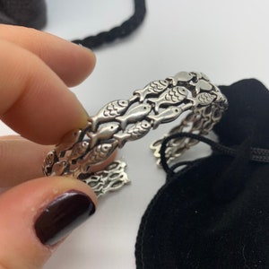 May include: A silver cuff bracelet featuring a repeating pattern of fish. The bracelet is open at one end and appears to be made of a metal alloy. The fish are detailed with fins and scales. A black drawstring bag is in the background.