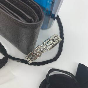 May include: A silver-toned ring featuring a repeating pattern of fish. The ring is set against a white background, with a black wallet, a blue bottle, and a black rope in the background.