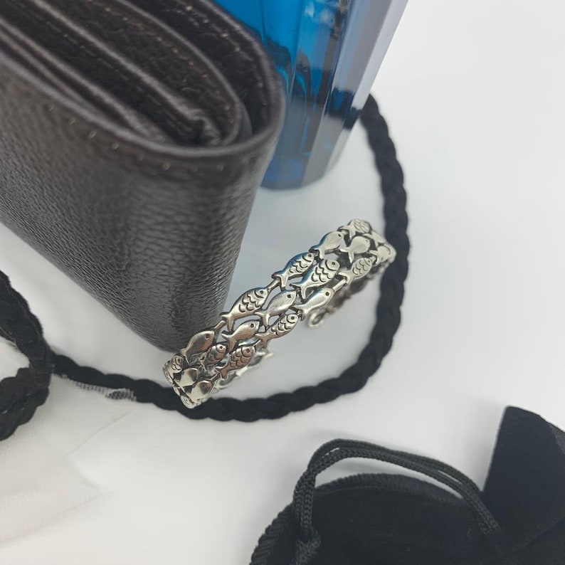 May include: A silver-toned ring featuring a repeating pattern of fish. The ring is set against a white background, with a black wallet, a blue bottle, and a black rope in the background.