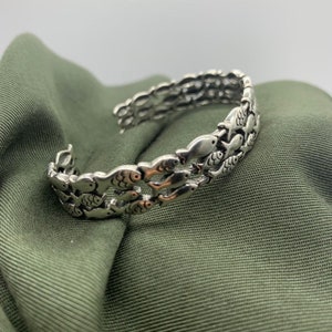 May include: Silver-toned cuff bracelet featuring a repeating pattern of fish. The bracelet has an open design and is resting on a green fabric surface. The fish are detailed with eyes and fins.