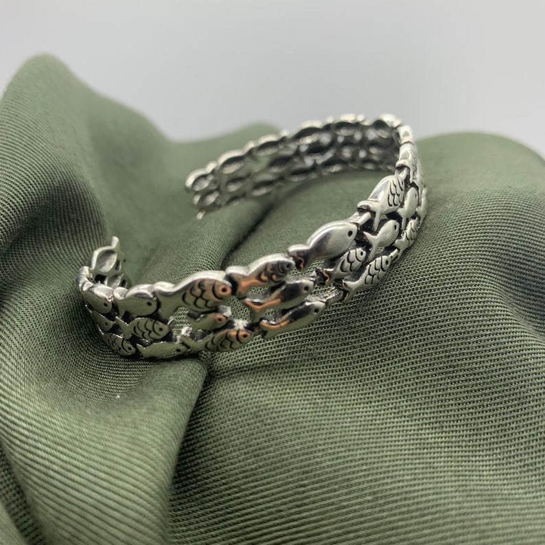 May include: Silver-toned cuff bracelet featuring a repeating pattern of fish. The bracelet has an open design and is resting on a green fabric surface. The fish are detailed with eyes and fins.