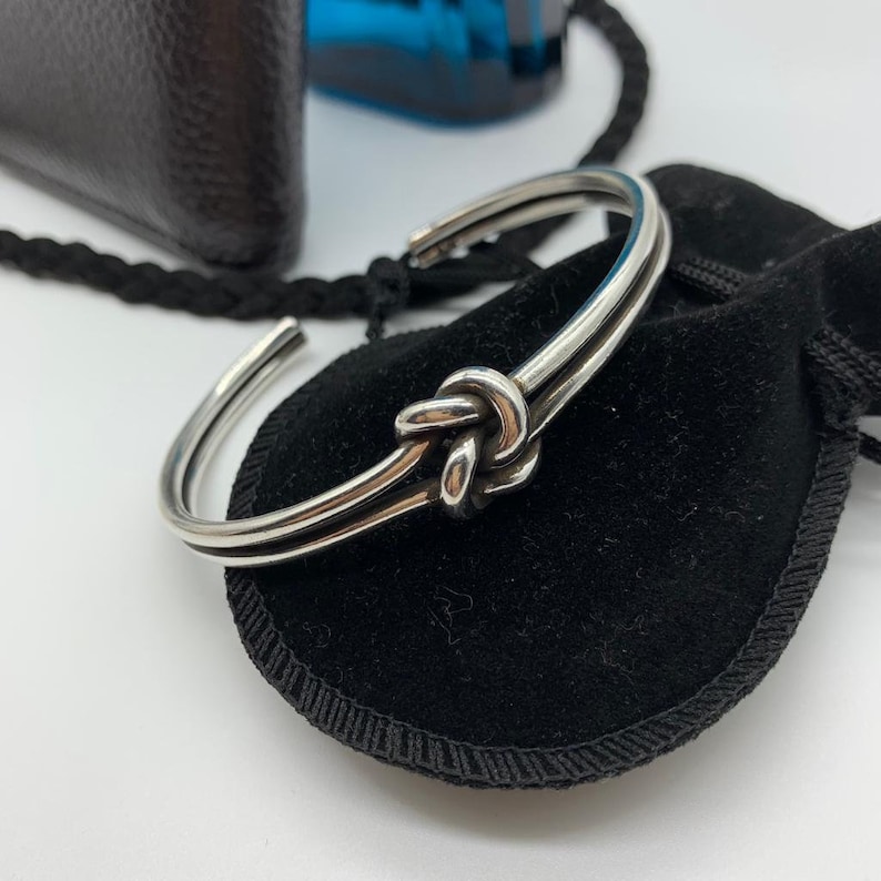 May include: A silver knot bracelet with a double band design. The bracelet is on a black velvet pouch.