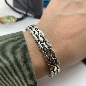 May include: Silver bracelet featuring a repeating fish design. The bracelet is made of multiple rows of fish, creating a unique and eye-catching accessory. The bracelet is worn on a wrist, showcasing its intricate details.