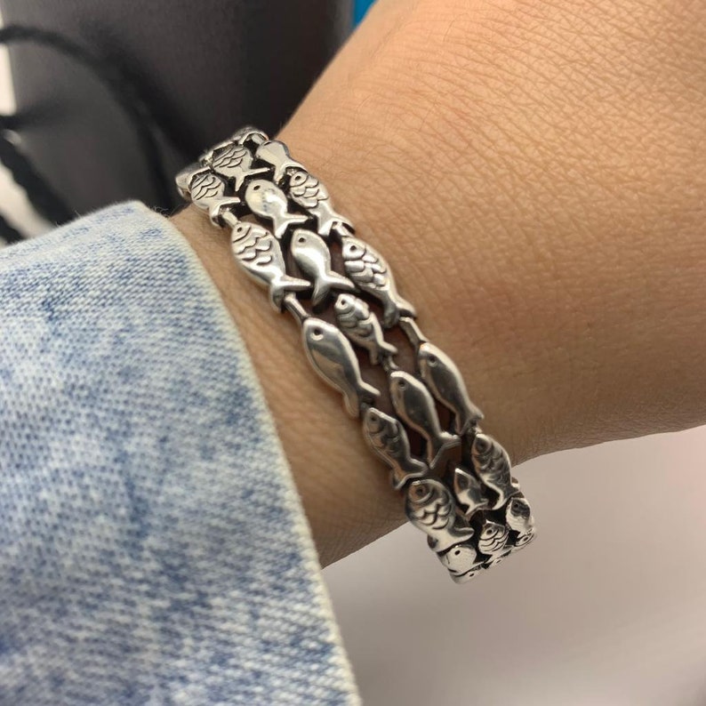 May include: A silver bracelet featuring a repeating pattern of fish. The bracelet is made of multiple rows of fish-shaped links, creating a textured and detailed design. The bracelet is worn on a wrist.