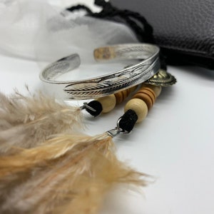 May include: A silver bracelet with a feather design, wooden beads, and a feather charm. The bracelet is on a white surface with a white and black mesh bag in the background.