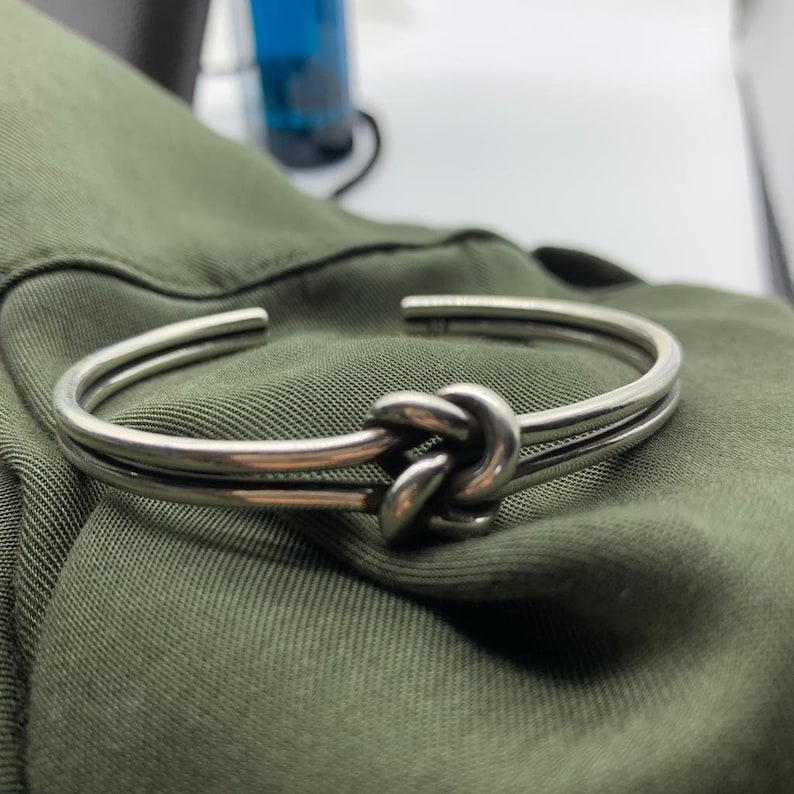 May include: A silver knot bracelet with a double band design. The bracelet is made of polished metal and has a simple, elegant style.