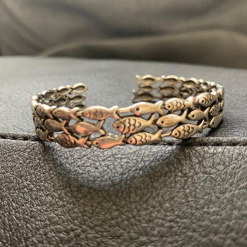 May include: Silver-toned cuff bracelet featuring a repeating pattern of fish. The bracelet has an open design, allowing it to be easily put on and taken off. The fish are detailed with fins and eyes, creating a nautical theme.