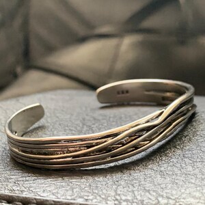 May include: A silver bracelet with a woven design. The bracelet is open and has a simple, minimalist style.