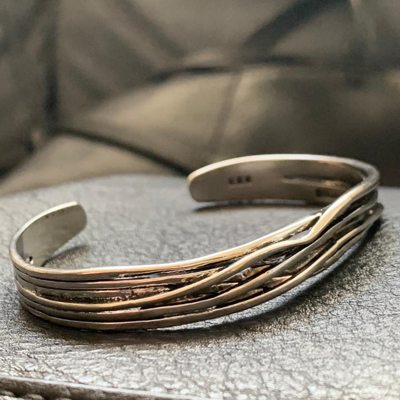 May include: A silver bracelet with a woven design. The bracelet is open and has a simple, minimalist style.