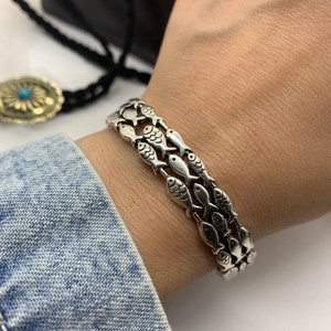 May include: A silver bracelet featuring a repeating fish design. The bracelet is composed of three rows of fish, each meticulously detailed. The bracelet is worn on a wrist, showcasing its intricate craftsmanship and aquatic theme.