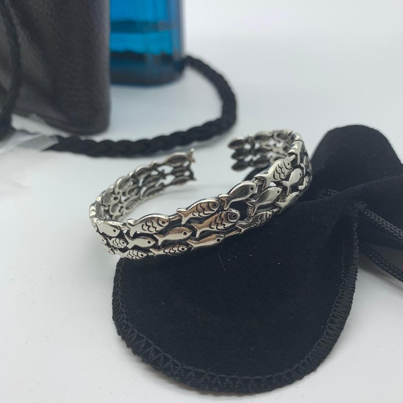 May include: A silver cuff bracelet featuring a repeating pattern of small fish. The open-ended bracelet is displayed on a black velvet pouch. The bracelet has a nautical theme and is likely made of sterling silver.