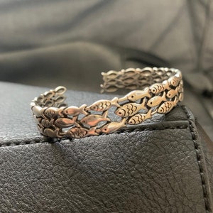 May include: Silver-toned cuff bracelet featuring a repeating pattern of small fish. The open-ended bracelet has a textured surface and is designed to be worn on the wrist. The fish are arranged in rows, creating a unique aquatic design.