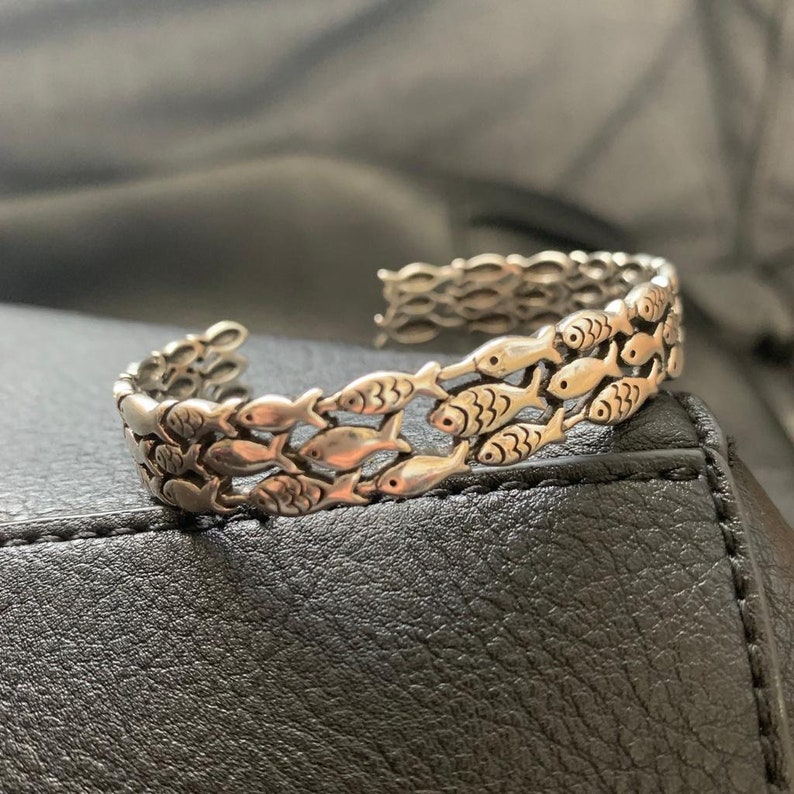 May include: Silver-toned cuff bracelet featuring a repeating pattern of small fish. The open-ended bracelet has a textured surface and is designed to be worn on the wrist. The fish are arranged in rows, creating a unique aquatic design.