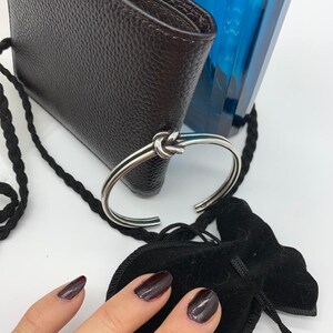 May include: A silver bangle bracelet with a knotted design. The bracelet is lying on a brown leather wallet and a black velvet pouch.