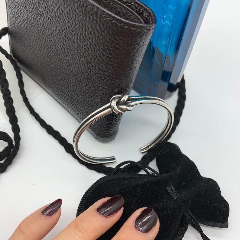 May include: A silver bangle bracelet with a knotted design. The bracelet is lying on a brown leather wallet and a black velvet pouch.
