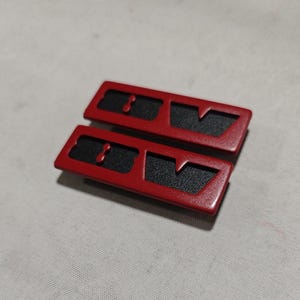 Puede incluir: Dos molduras rectangulares rojas para coche con inserciones negras texturizadas. Cada pieza tiene un diseño recortado único y un pequeño detalle circular. Los artículos se muestran sobre una superficie gris claro.