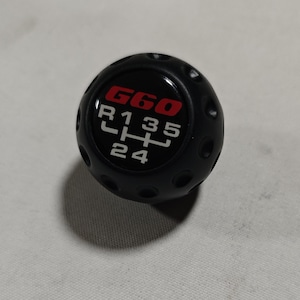Könnte beinhalten: Schwarzer Schalthebelknauf mit dem roten Text "G60" und dem weißen Gangmuster. Der Knauf hat eine runde Form mit kleinen kreisförmigen Vertiefungen an den Seiten.