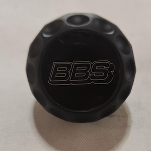 Op de afbeelding: Een zwarte wielnaafdop met een verhoogd, getextureerd oppervlak en het woord "BBS" in witte letters.
