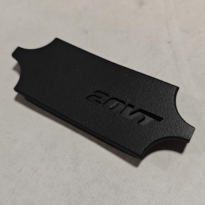 Peut inclure: Un emblème rectangulaire noir mat avec une surface texturée et des côtés crantés. L'emblème présente le texte 20VT en relief dans une police angulaire stylisée. L'objet est rigide et possède une forme géométrique nette aux bords définis.