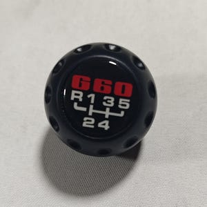 Può includere: Pomello del cambio nero con la scritta "G60" in rosso e un schema del cambio bianco. Il pomello ha piccoli fori tutt'intorno. Lo schema include "R, 1, 2, 3, 4, 5".