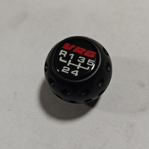 Könnte beinhalten: Schwarzer Schalthebelknauf mit dem Text "VR6" in Rot und Gangmuster-Markierungen in Weiß. Der Knauf hat eine kugelförmige Form mit kleinen kreisförmigen Vertiefungen.