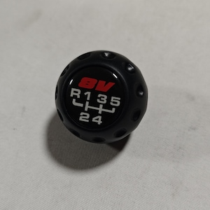 Könnte beinhalten: Schwarzer Schalthebelknauf mit kreisförmigem Design. Der Knauf hat ein rot-weißes Gangmuster mit den Buchstaben und Zahlen: BV, R, 1, 3, 5, H, 2 und 4. Der Knauf hat kleine kreisförmige Vertiefungen an den Seiten.