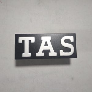 Könnte beinhalten: Ein schwarzes rechteckiges Schild mit den weißen Buchstaben „TAS“ in fetter, erhabener Schrift. Das Schild ist mittig platziert und scheint aus Holz oder einem ähnlichen Material zu bestehen. Der Hintergrund ist eine schlichte, hellgraue Oberfläche.