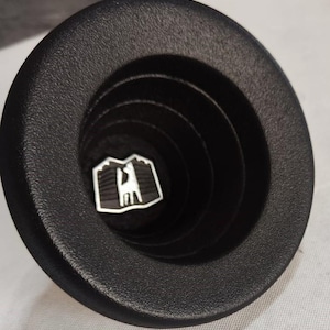 May include: A black car vent with a circular design and a textured finish. The vent features concentric rings and a central emblem. The emblem is a white and black graphic of a mountain and a stylized figure.