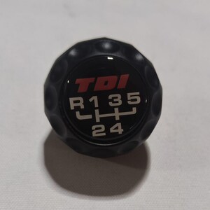 May include: Black gear shift knob featuring the text "TDI" in red and white. The shift pattern is shown with the numbers R, 1, 3, 5, 2, and 4. The knob has a textured, rounded design.