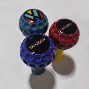 Op de afbeelding: Drie golfbalvormige knoppen met verschillende kleuren en ontwerpen. De eerste knop is blauw met een wit en geel Volkswagen-logo. De tweede knop is rood met het woord "HARLEQUIN" in witte letters. De derde knop is blauw met het woord "HARLEQUIN" in regenboogkleuren.