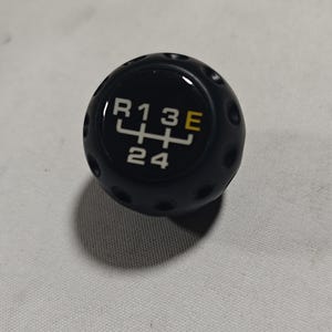 May include: Black gear shift knob with white and yellow markings. The knob has a diagram of the gear positions: R, 1, 3, E, 2, and 4. The knob has small dimples around the sides.